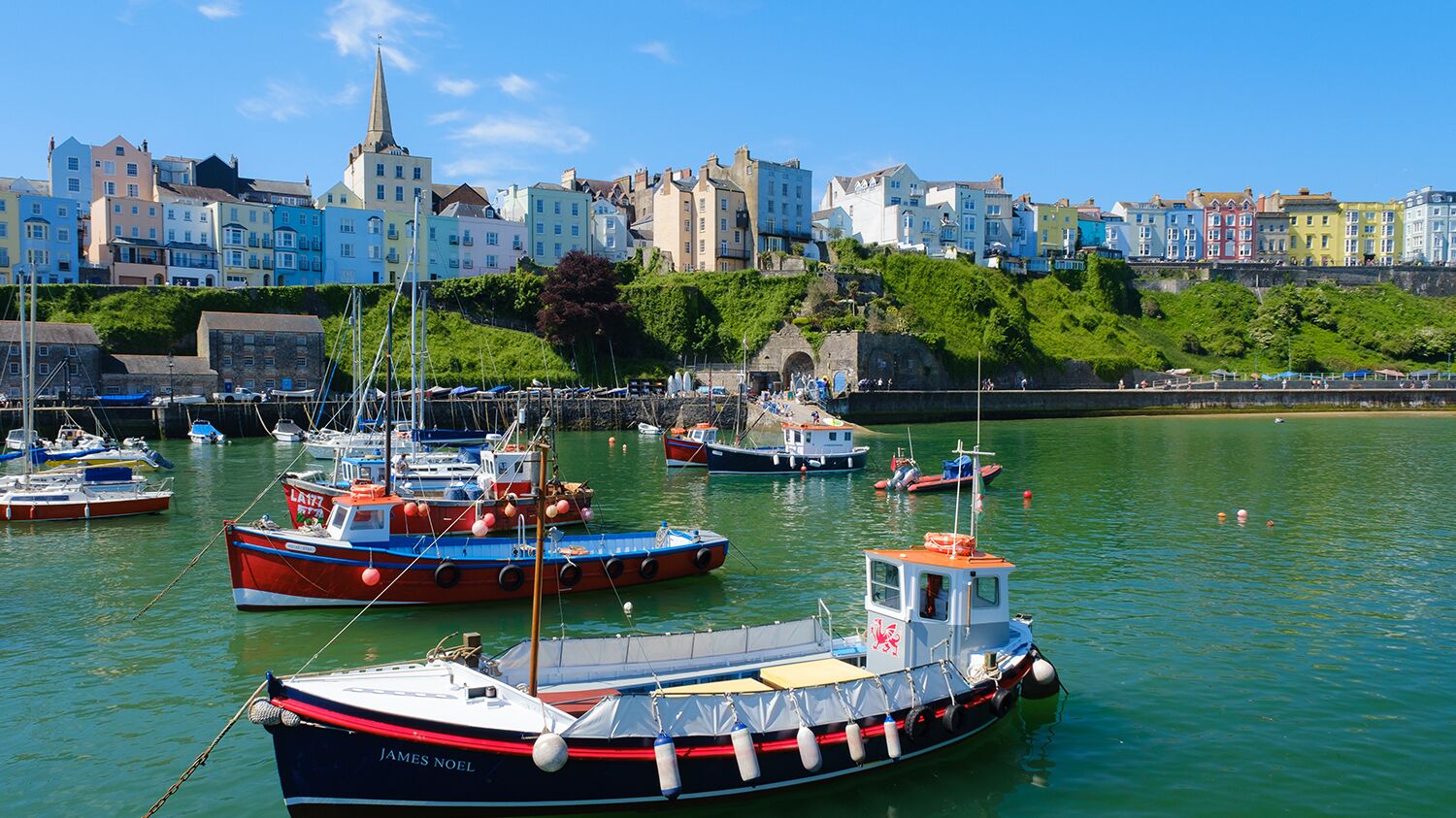 Un port où sont amarrés des bateaux, le clocher d’une église et des bâtiments aux couleurs vives qui bordent le front de mer.