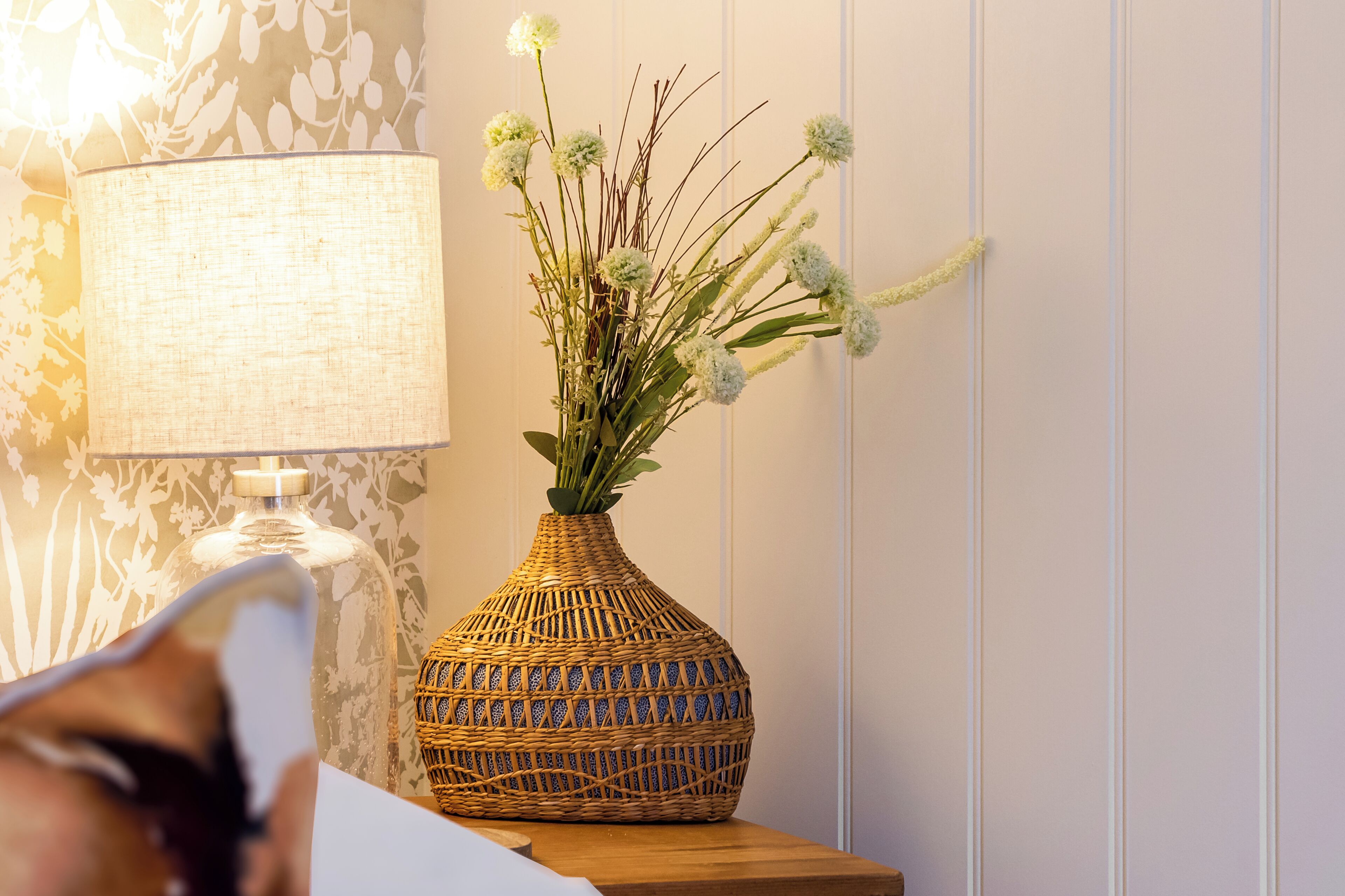 Un florero de mimbre con un arreglo floral sobre una mesa de madera junto a una lámpara.