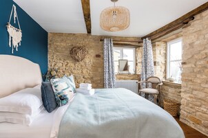 Un dormitorio con pared de piedra, cama con ropa blanca, decoración de macramé colgante y lámpara de mimbre.