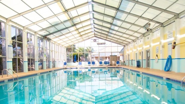A large indoor swimming pool with clear blue water, surrounded by lounge chairs and a covered roof structure.