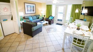 A compact living space with a sofa, a dining table set for two, a TV mounted on the wall, and a washing machine in the corner.