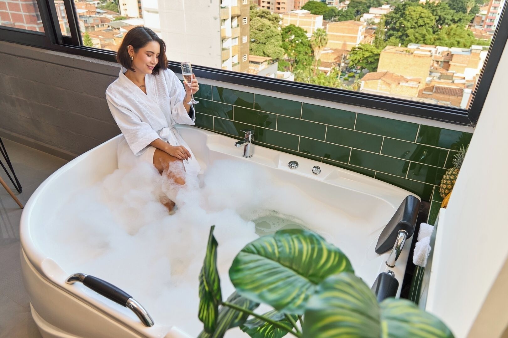 Une personne vêtue d’un peignoir blanc est assise dans une grande baignoire remplie de mousse, et on peut apercevoir des bâtiments et de la verdure à travers la fenêtre.