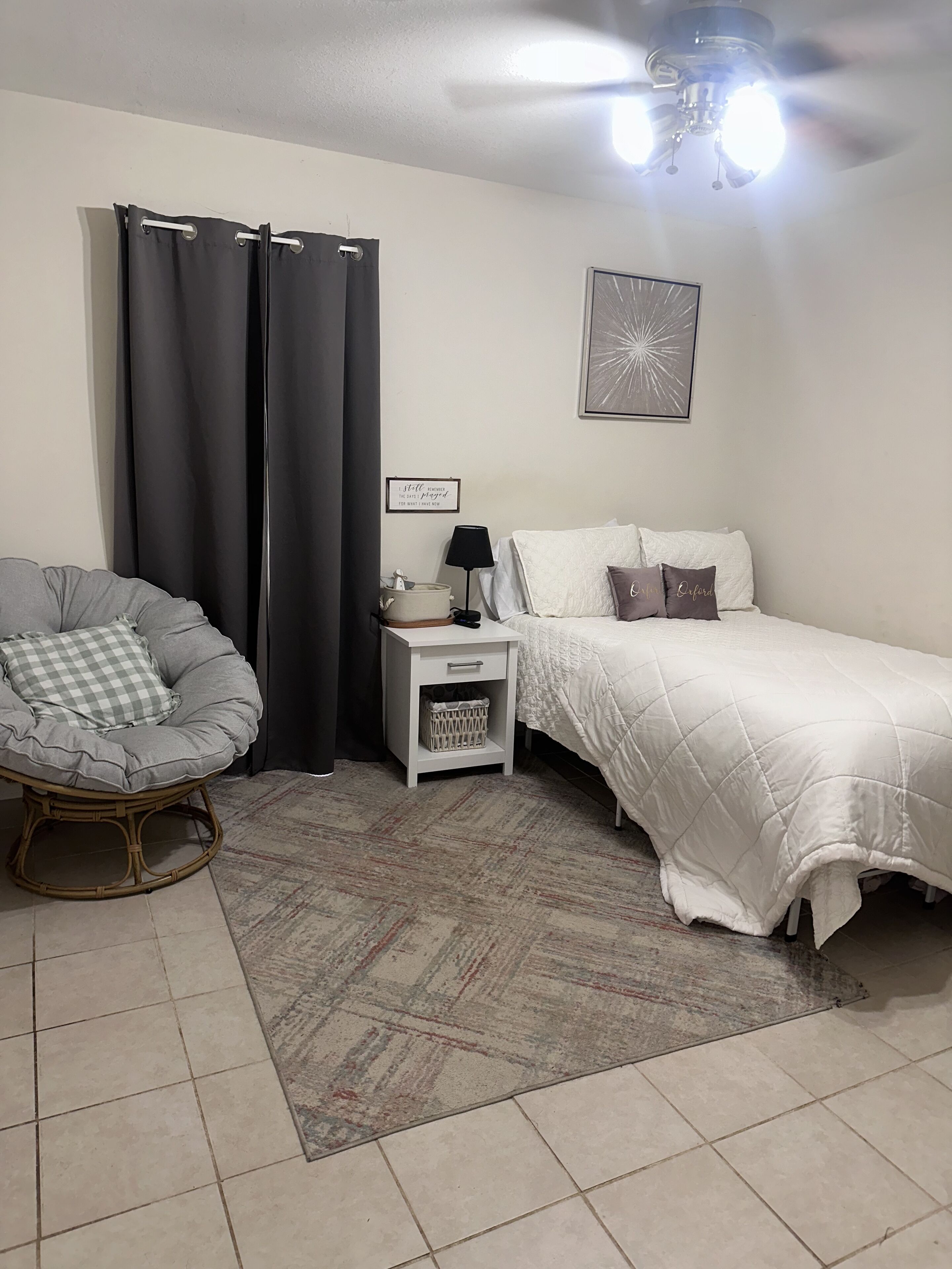 Une chambre à coucher comprenant un lit, une chaise, une table de chevet, une lampe, un ventilateur de plafond et un tapis.