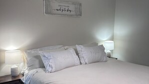 Ein ordentlich bezogenes Bett mit weißen Leinen und Kissen, eine Nachttischlampe und eine Wandleuchte.
