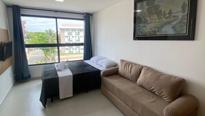 Ein Hotelzimmer mit einem Bett, einem beigefarbenen Sofa, einem großen Fenster mit Vorhängen, einem Bild an der Wand und einem gefliesten Boden.
