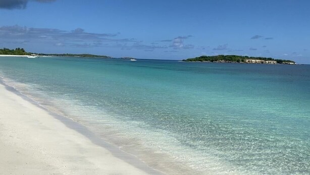 Una playa con aguas turquesas cristalinas, una orilla arenosa y una pequeña isla con vegetación al fondo.