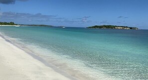 Ein Strand mit klarem türkisblauem Wasser, einem sandigen Ufer und einer kleinen Insel mit Vegetation in der Ferne.