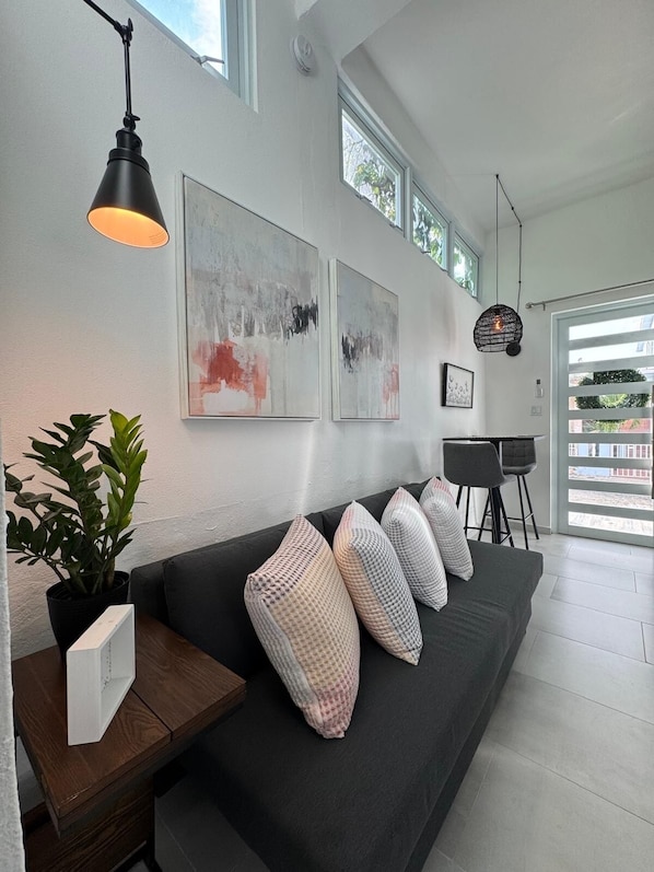 A modern living room with a dark grey sofa, abstract wall art, a potted plant, and a dining area with a black chair and table.