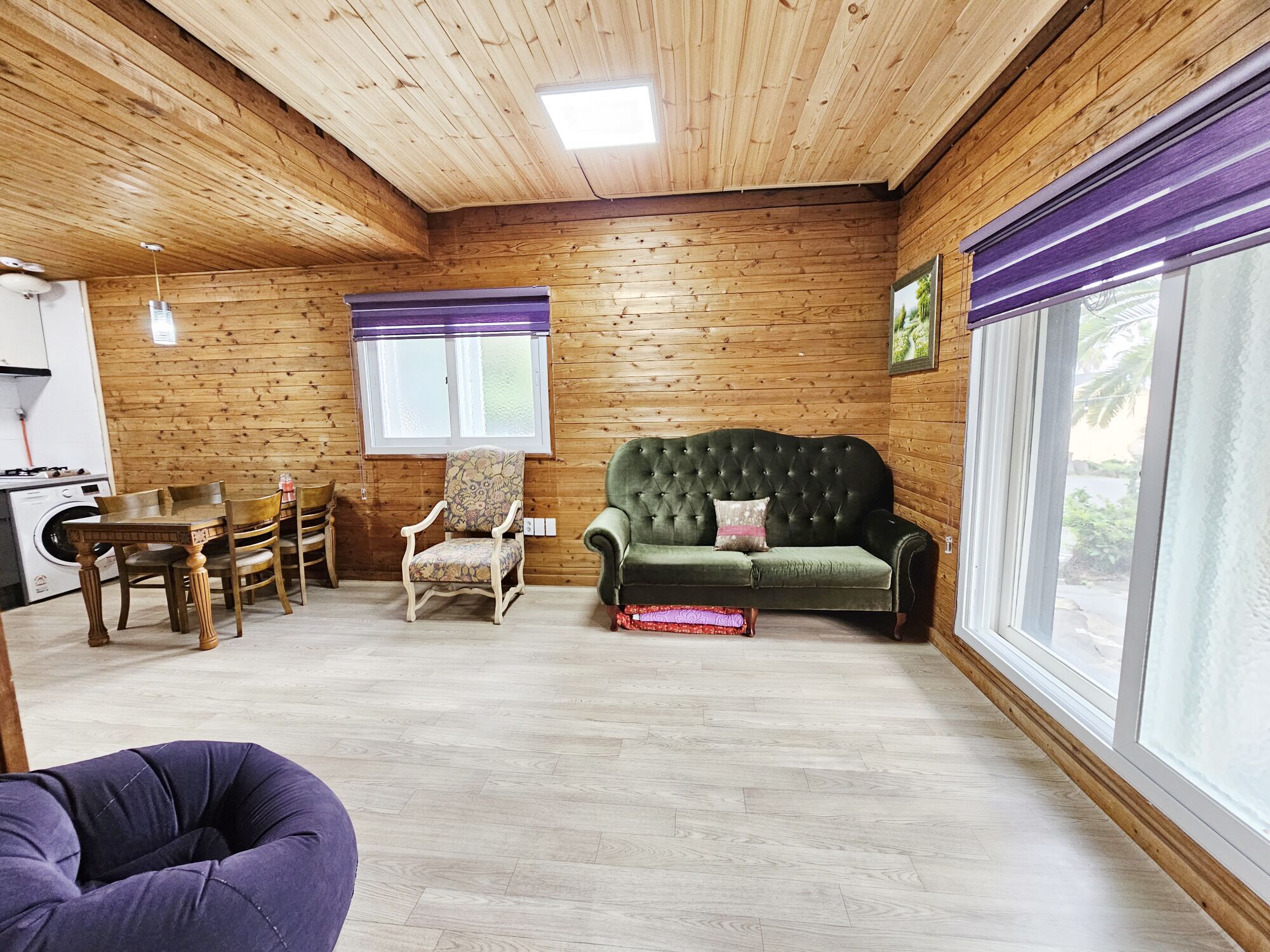 A cozy room with wooden walls, a green sofa, a patterned armchair, a dining table with chairs, and a large window with a view.