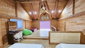 A wooden cabin room with two beds, a green bean bag, a desk with a computer, and a window with blinds. - Tree Story Pension (Seogwipo)