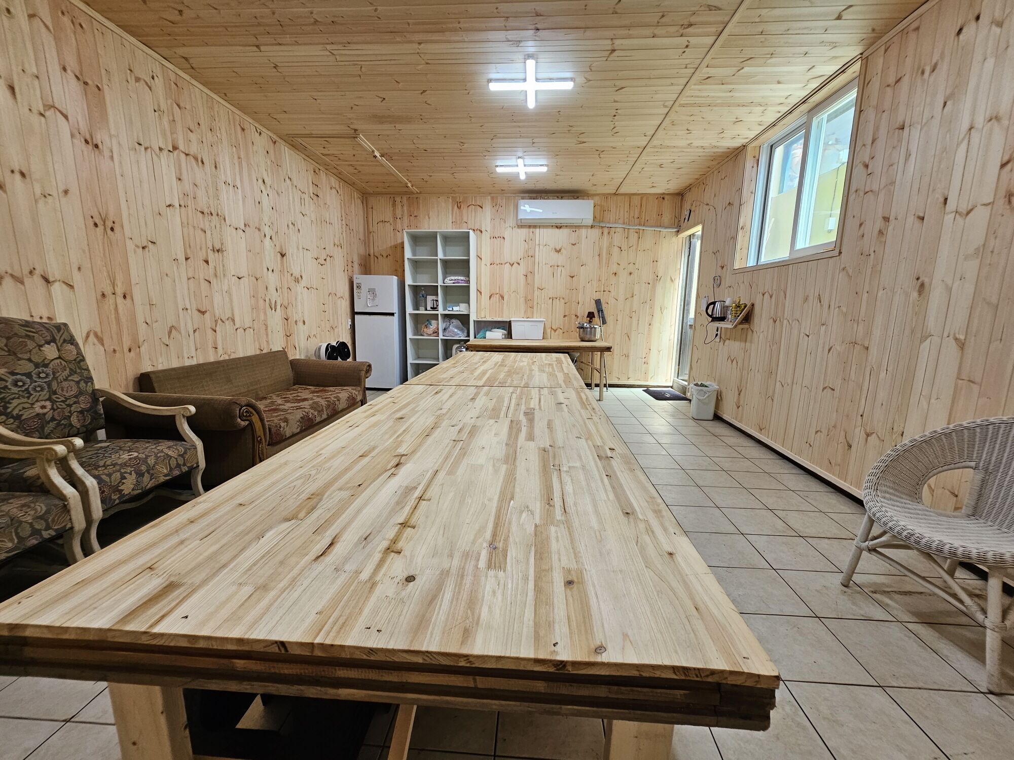 A wooden-paneled room with a long table, two sofas, a wicker chair, and a kitchen area with a refrigerator and shelves.