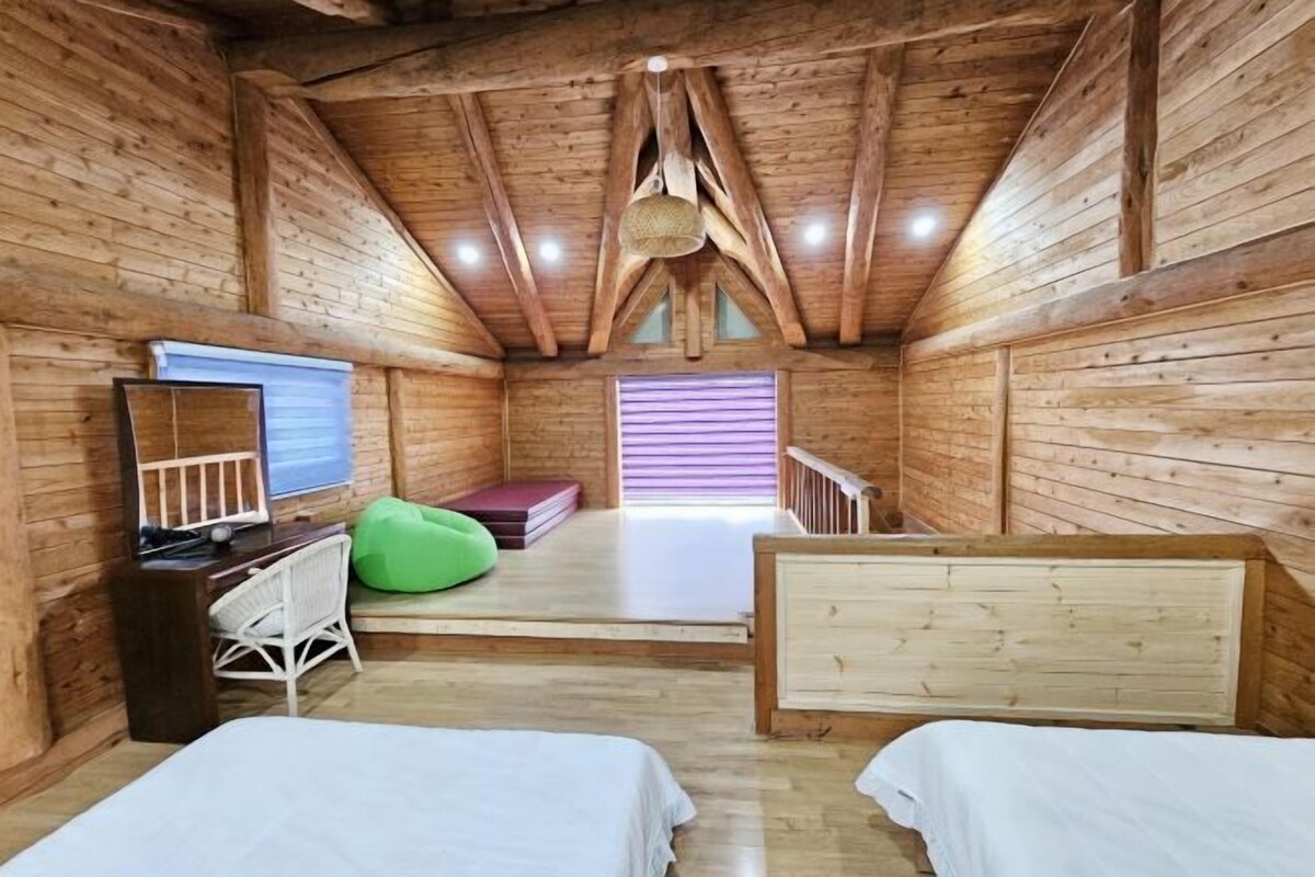Ein gemütliches Blockhauskabinenzimmer mit Holzwänden, einem Schlafbett im Zwischengeschoss, einem Schreibtisch mit Computer, einem grünen Sitzsack und einem Fenster mit Jalousien.