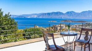 Una terraza con dos sillas de madera y una mesa redonda pequeña, con vista al mar azul y a las montañas.