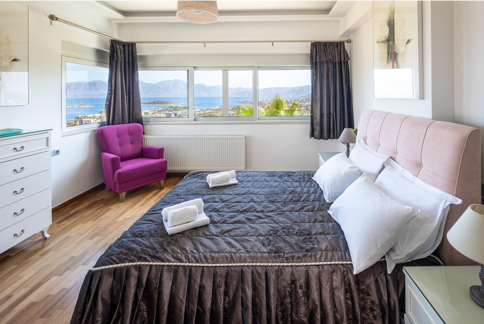 A bedroom with a bed, a chair, a dresser, and a view of mountains and sea.