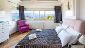 Un dormitorio con cama, una silla, tocador y vistas a las montañas y al mar.