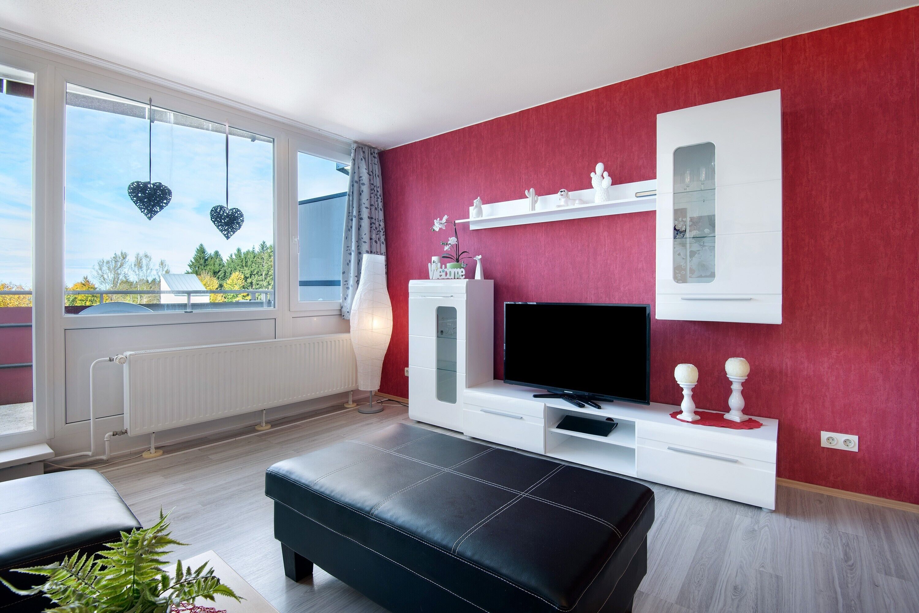 Una sala de estar moderna con una pared de acento roja, un mueble blanco para entretenimiento, un sofá de cuero negro y un ventanal con vista a árboles.