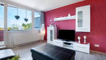 Una sala de estar moderna con una pared de acento roja, un mueble blanco para entretenimiento, un sofá de cuero negro y un ventanal con vista a árboles.