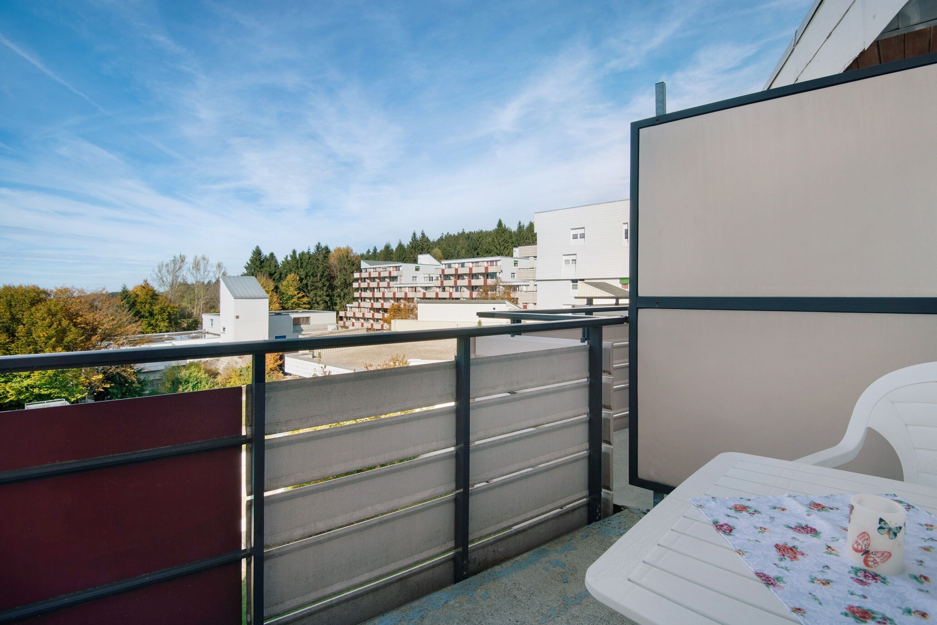 Un balcone con una sedia bianca, una ringhiera in metallo e vista su edifici e alberi.
