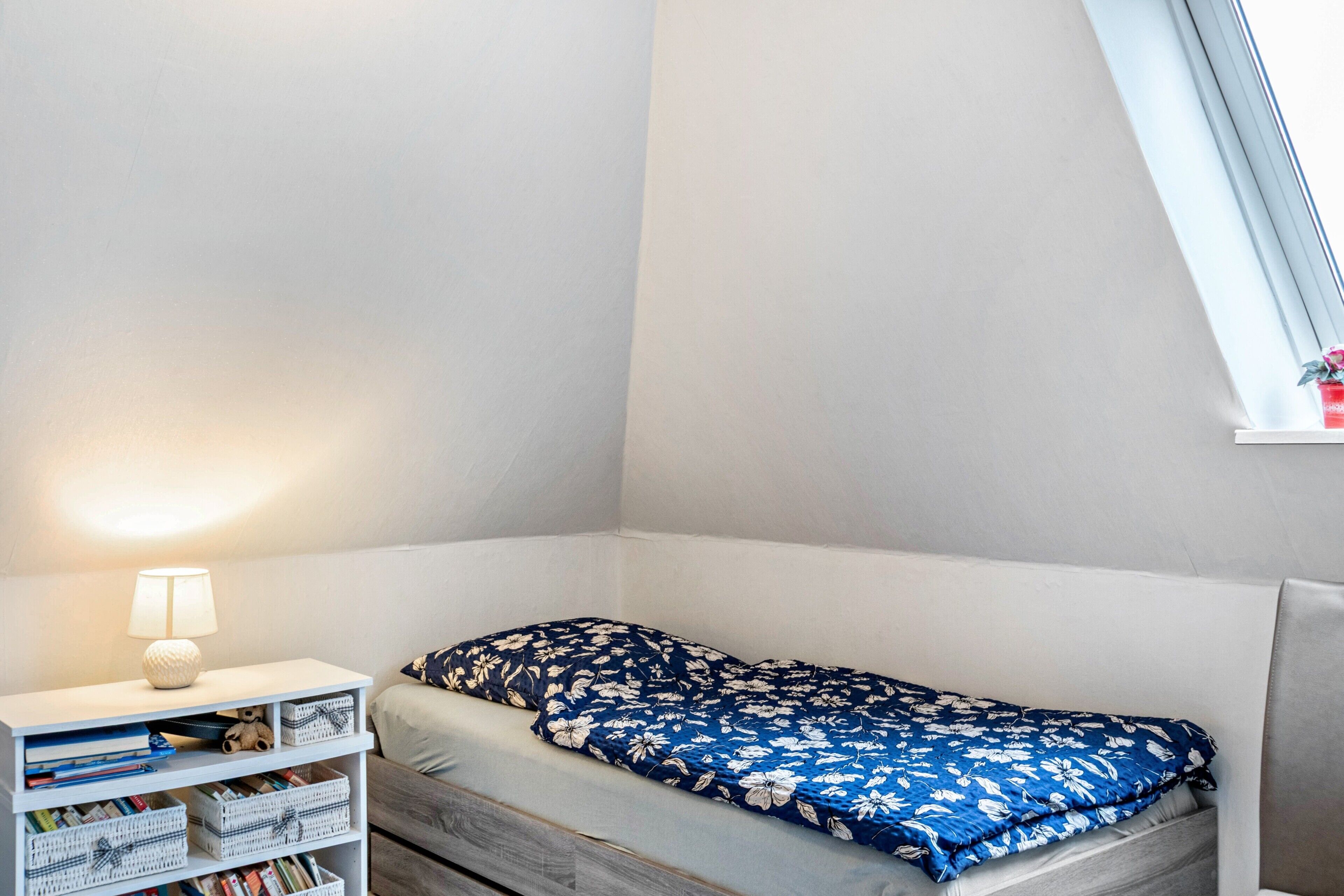 Une petite chambre à coucher, bien rangée, avec un lit simple, une table de chevet avec une lampe et une fenêtre donnant sur un beau paysage.