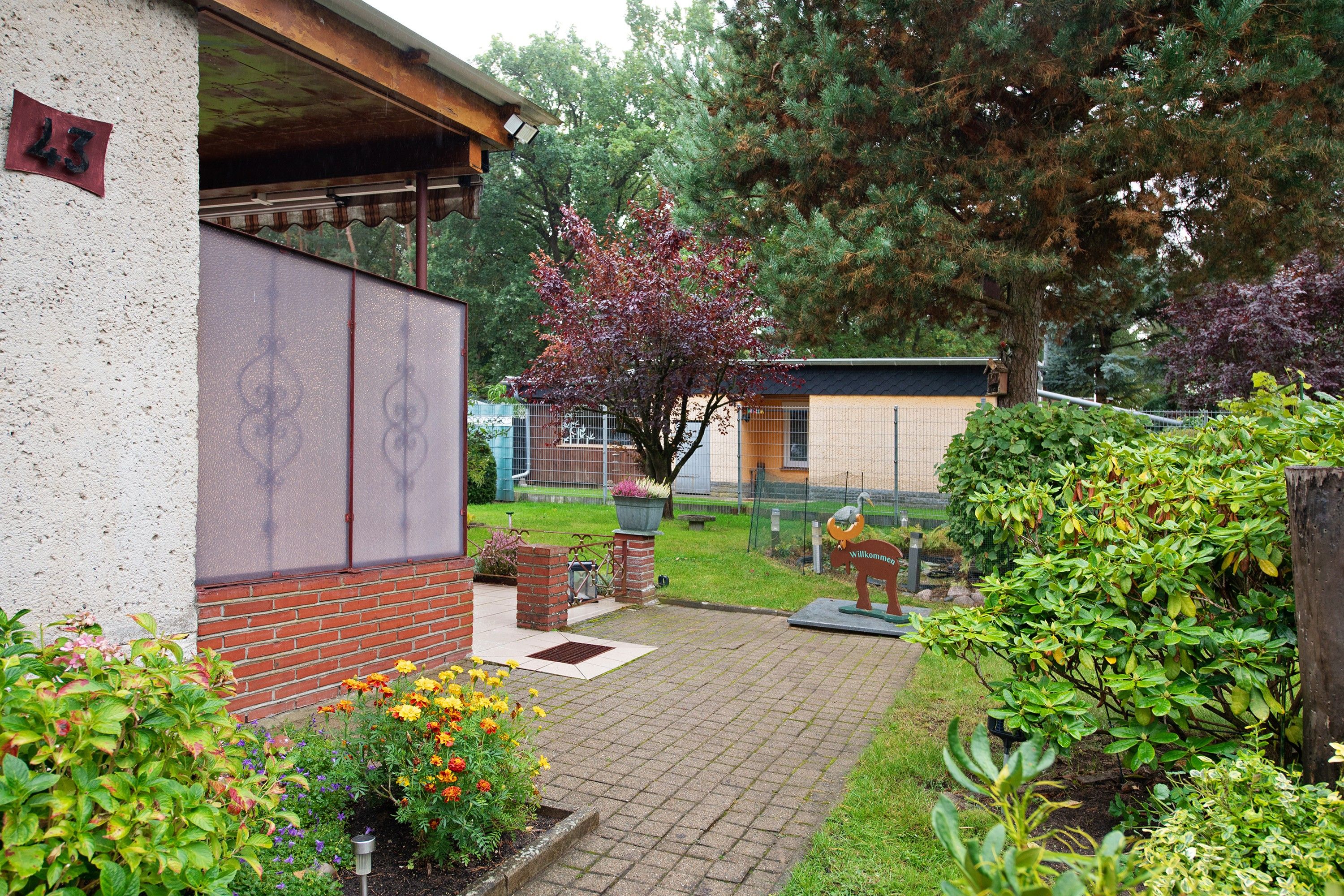 Una propiedad residencial con un sendero pavimentado que conduce a una puerta de madera, rodeada de abundante follaje y diversas plantas.