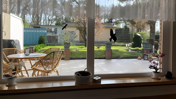 Eine Terrasse mit Korbmöbeln, einem Topfpflanzen und Blick auf einen Garten mit Zaun und Bäumen.
