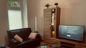 A living room with a black leather sofa, a glass-top coffee table, a flat-screen TV, and a wooden cabinet with plants.