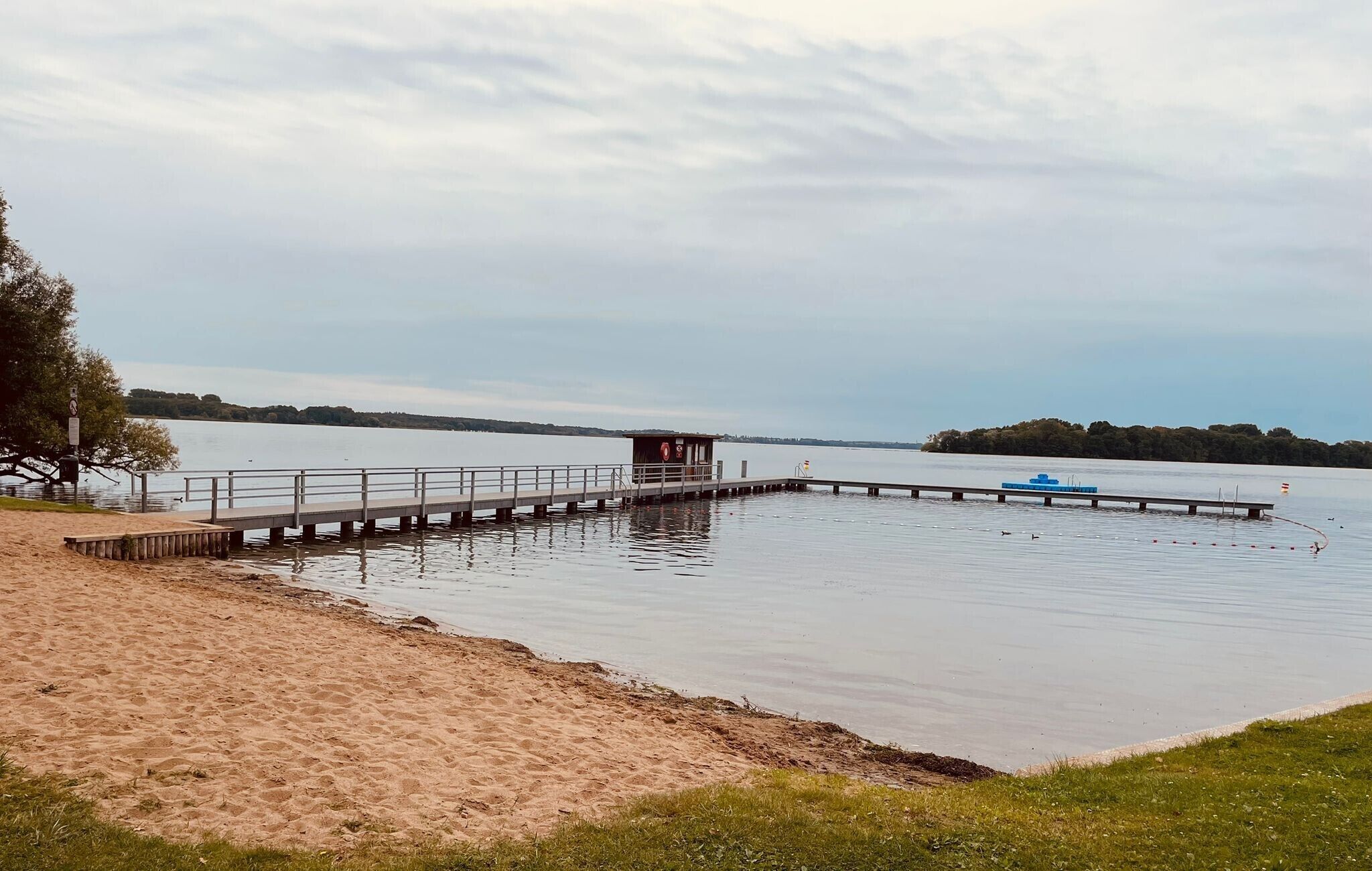 Una playa arenosa con un muelle de madera que se adentra en aguas tranquilas, una pequeña construcción de madera al final del muelle y una isla distante con árboles.