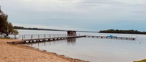 Una playa arenosa con un muelle de madera que se adentra en aguas tranquilas, una pequeña construcción de madera al final del muelle y una isla distante con árboles.