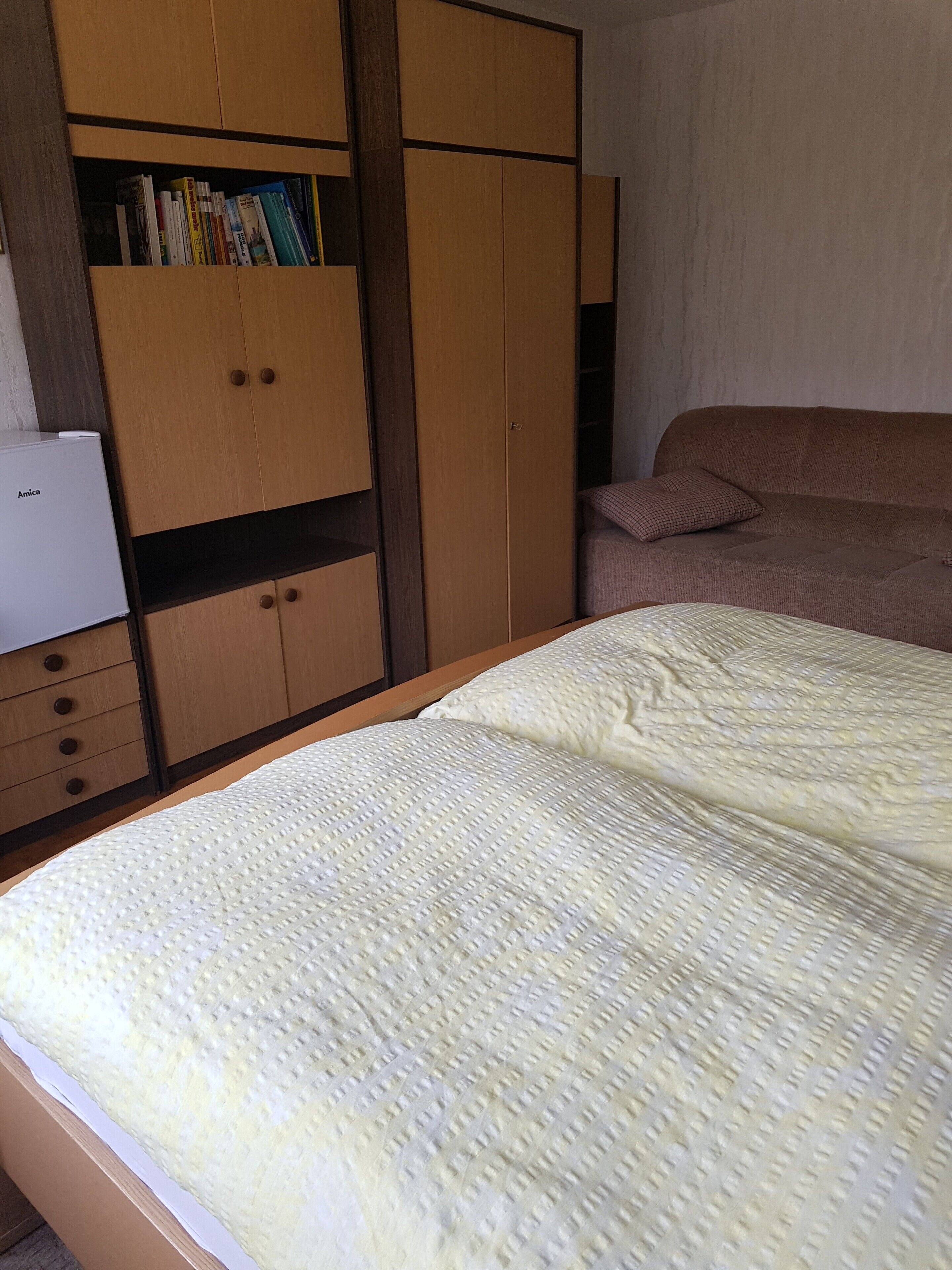 A bedroom with a bed, a wardrobe, and a small desk with a refrigerator.