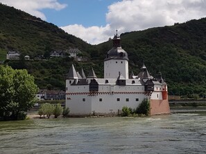 Ein schlossartiges Gebäude mit mehreren Türmen und einer zentralen Kuppel, umgeben von einem Fluss und üppigen grünen Hügeln.
