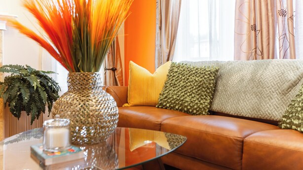 A living room with a brown leather sofa, a glass coffee table, a decorative vase with tall orange plants, and a green fern. - Celest Homes! Spacious, family home, right  for your next family adventure.  (Edmonton)