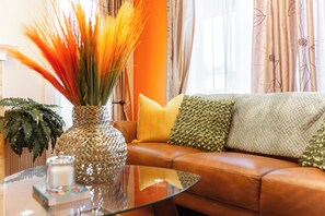 A living room with a brown leather sofa, a glass coffee table, a decorative vase with tall orange plants, and a green fern. - Celest Homes! Spacious, family home, right for your next family adventure. (Edmonton)