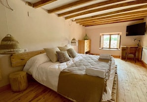 Un dormitorio con techo de madera, una cama con ropa blanca, una mesita de noche de madera, una lámpara colgante y una ventana con contraventanas.