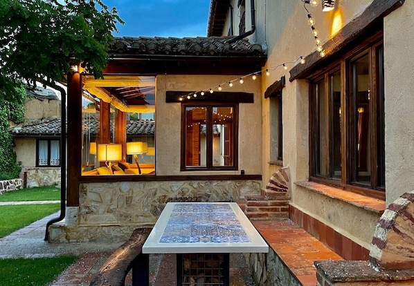 Un patio de piedra con una mesa de baldosas, un área de estar techada con lámparas y luces colgantes.