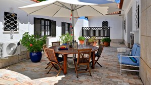 An outdoor patio with a round wooden table, chairs, a white umbrella, potted plants, and a white wall with a window and shutters.