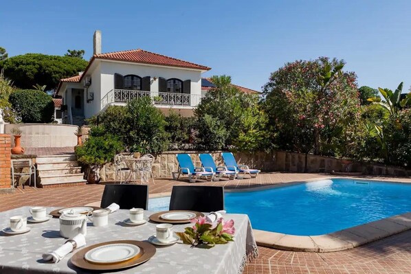 Un espace repas aménagé au bord de la piscine, avec une table dressée pour quatre personnes, une piscine et une maison dotée d’un balcon.