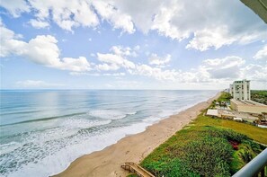 Vista da praia com céu claro, ondas e um edifício com vários andares ao fundo.