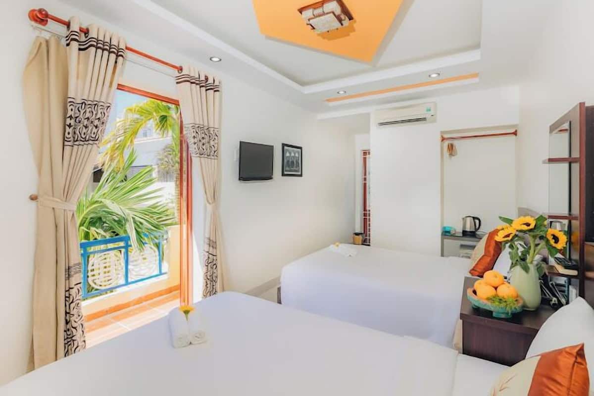 A hotel room with two beds, a large window with curtains, a ceiling light, an air conditioning unit, a flat-screen TV, and a small table with a vase of sunflowers.