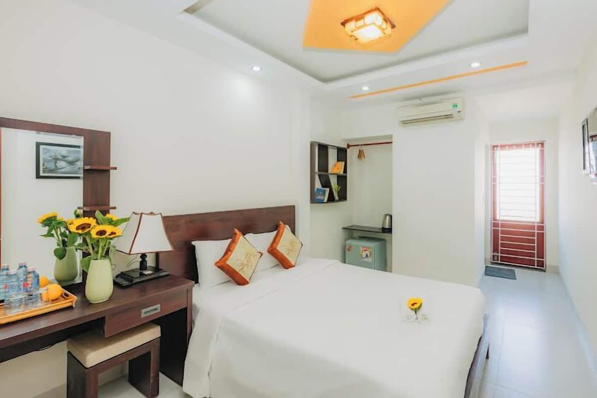 A hotel room with a bed, bedside table, lamp, and a small desk with a vase of sunflowers.