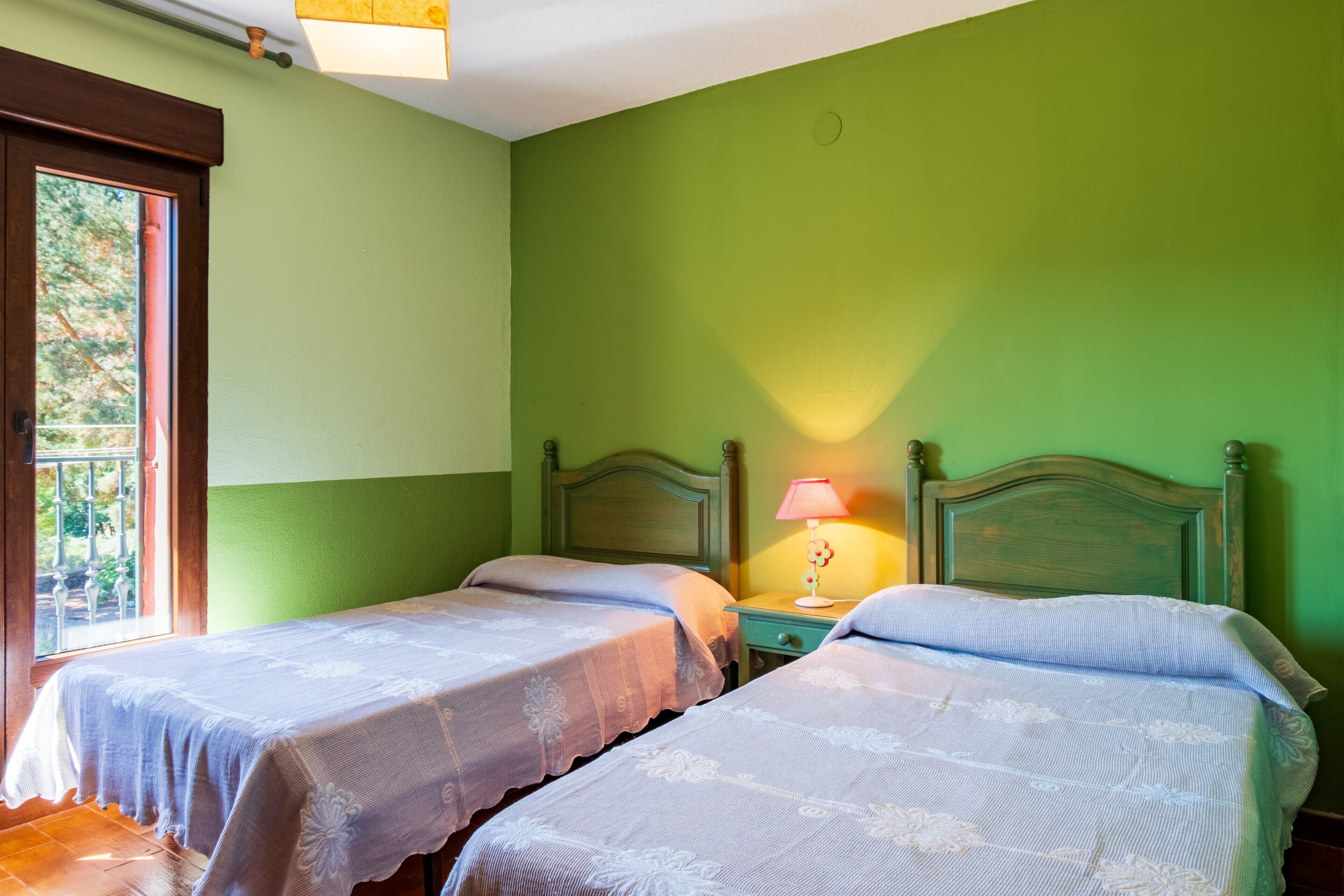 A room with two beds, a nightstand with a lamp, and a window with a view of trees.