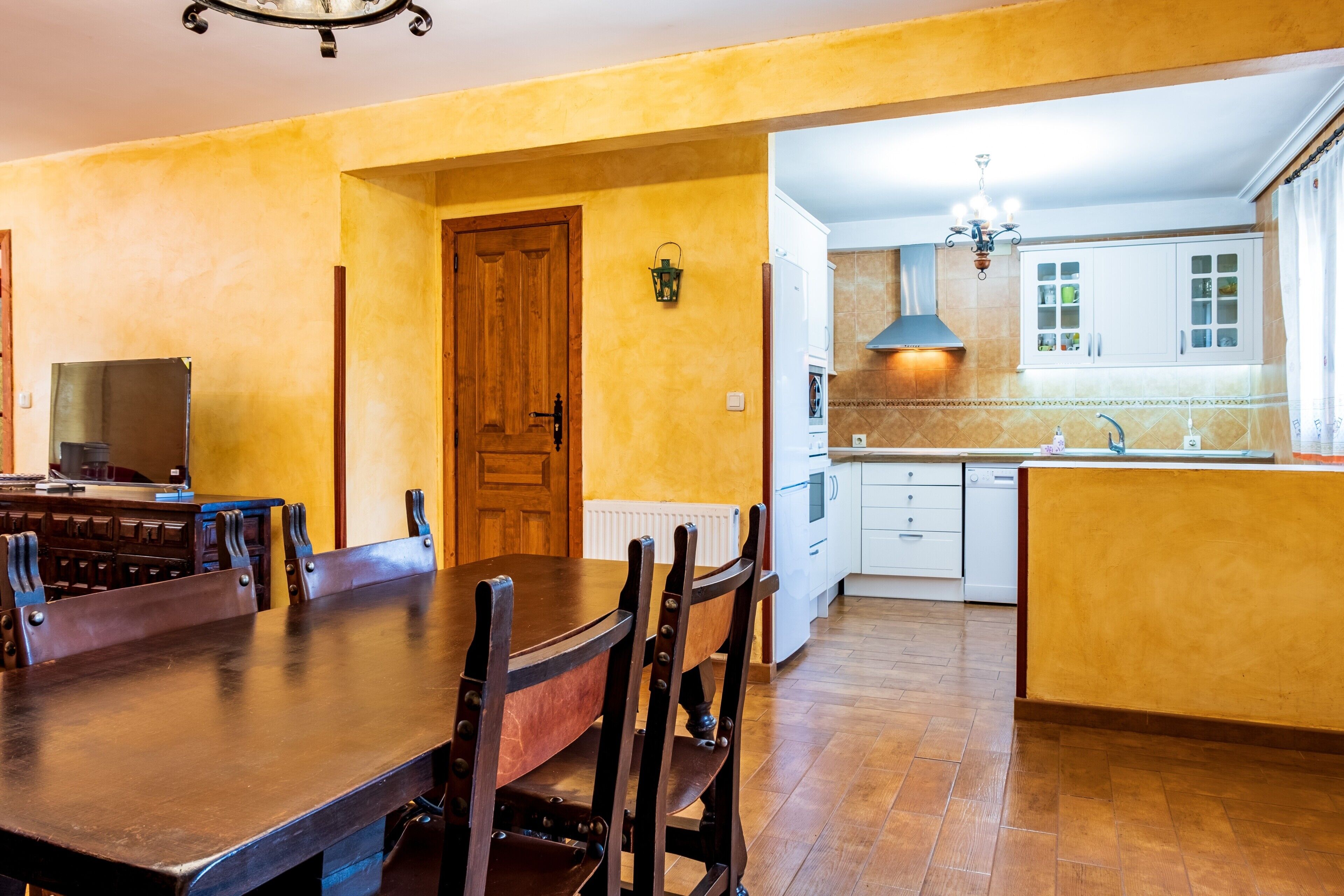 Comedor con mesa y sillas de madera, cocina con armarios blancos y puerta de madera.