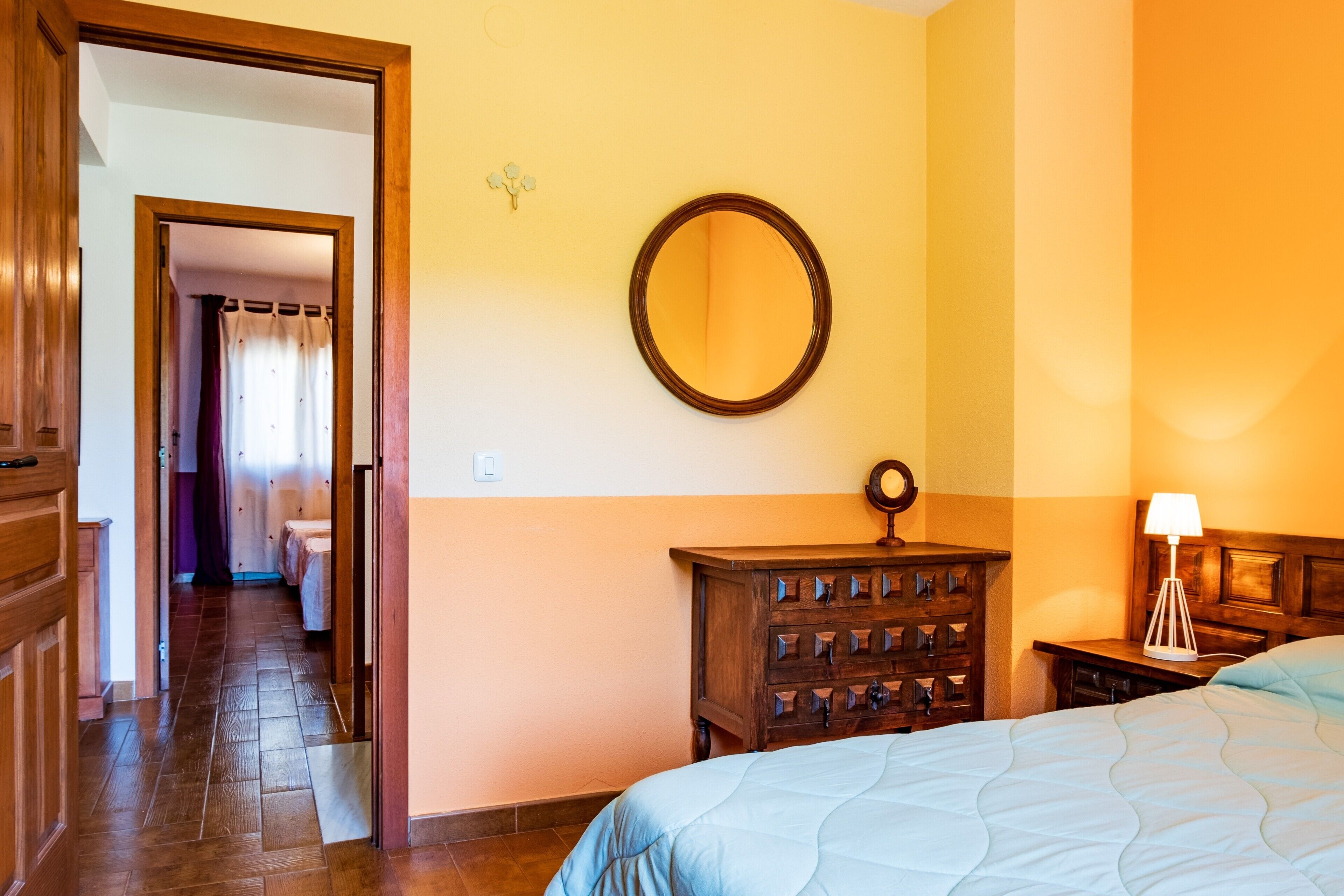A hotel room with a bed, a dresser, a nightstand, a mirror, and a door leading to another room.