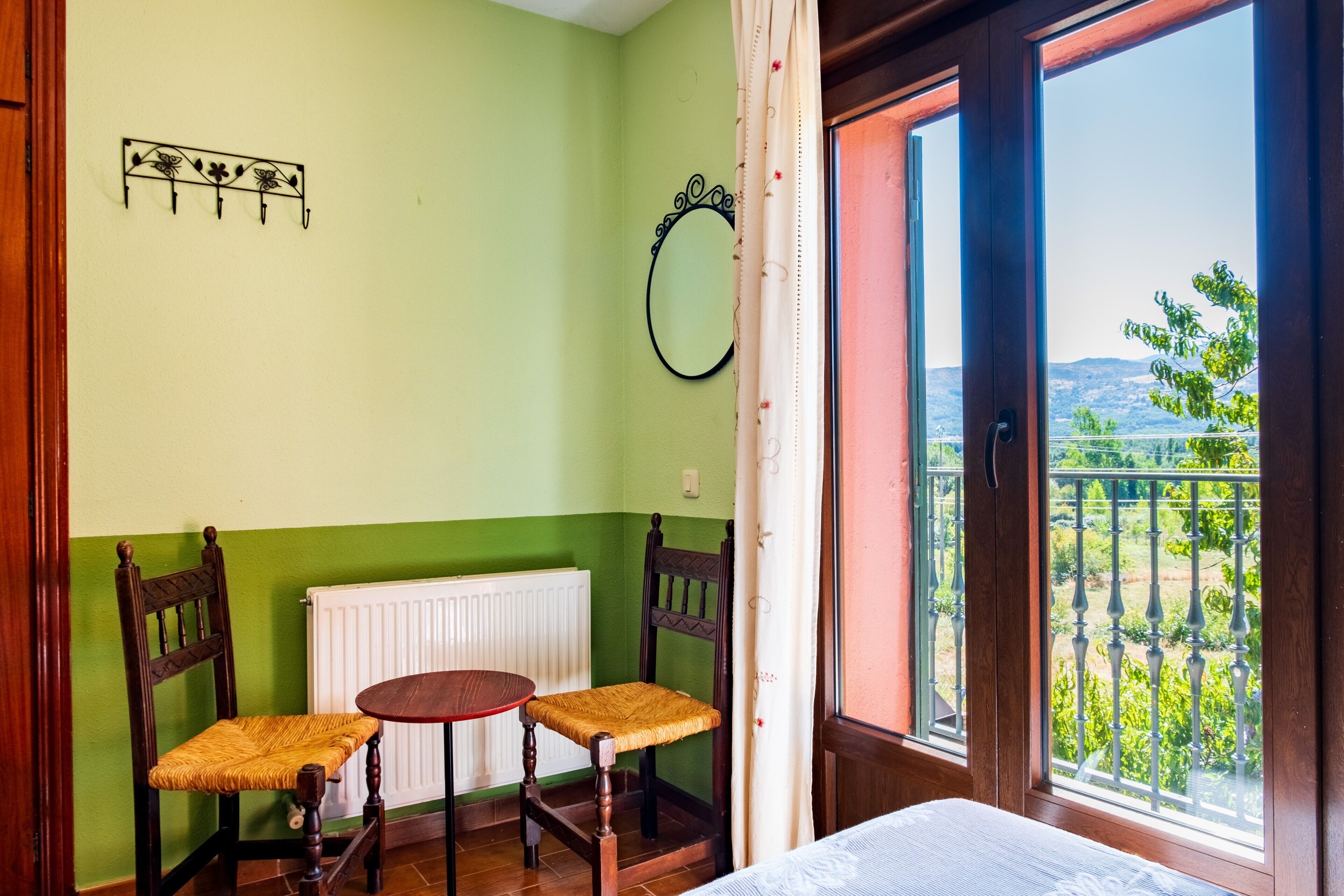 Una habitación con pared verde, una silla de madera, una mesa redonda pequeña, un espejo y vistas a la vegetación a través de una ventana.