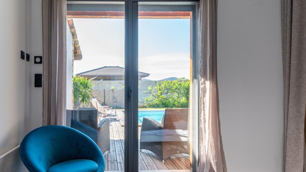 A room with a bed, a blue chair, and a sliding glass door leading to a pool area with outdoor furniture. - Holiday Home 'Villabergerie En Pierre Du Pays' with Private Pool, Wi-Fi and Air Conditioning (Barbaggio)