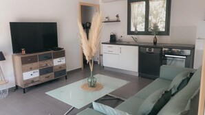 A modern living room with a flat-screen TV, a glass coffee table, a green sofa, a kitchen with a dishwasher, and a window with curtains.