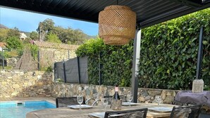 An outdoor patio with a wooden table, black wicker chairs, a hanging wicker light fixture, a stone wall, and a pool in the background.