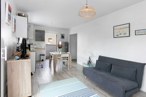 Uma sala moderna com um sofá cinzento, uma área de refeições com uma mesa de madeira e uma cozinha ao fundo.
