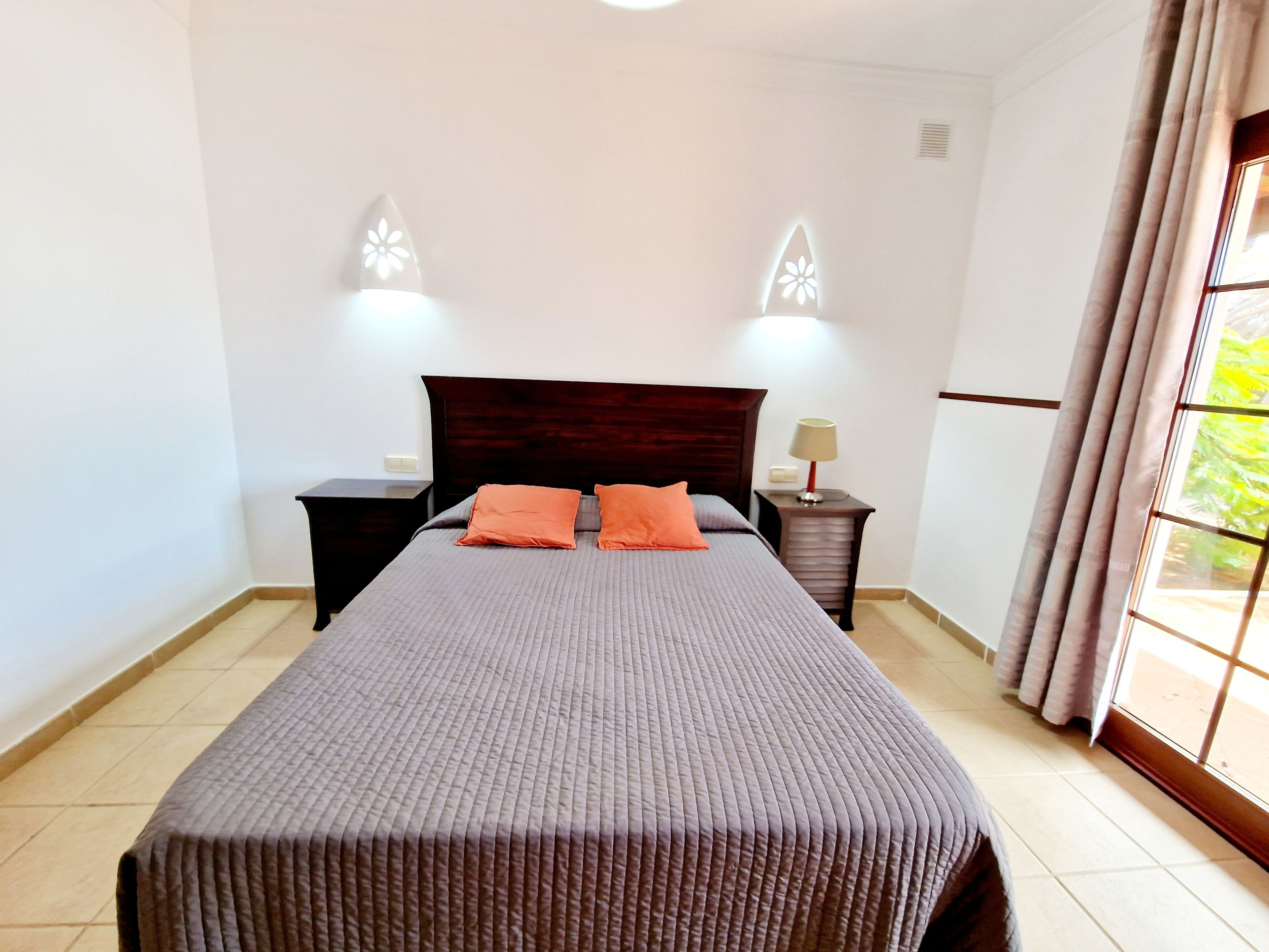 A neatly made bed with a dark headboard, two bedside tables with lamps, and a window with curtains.