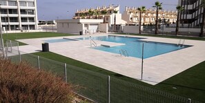 A swimming pool with a surrounding deck, a fenced area, and buildings in the background.
