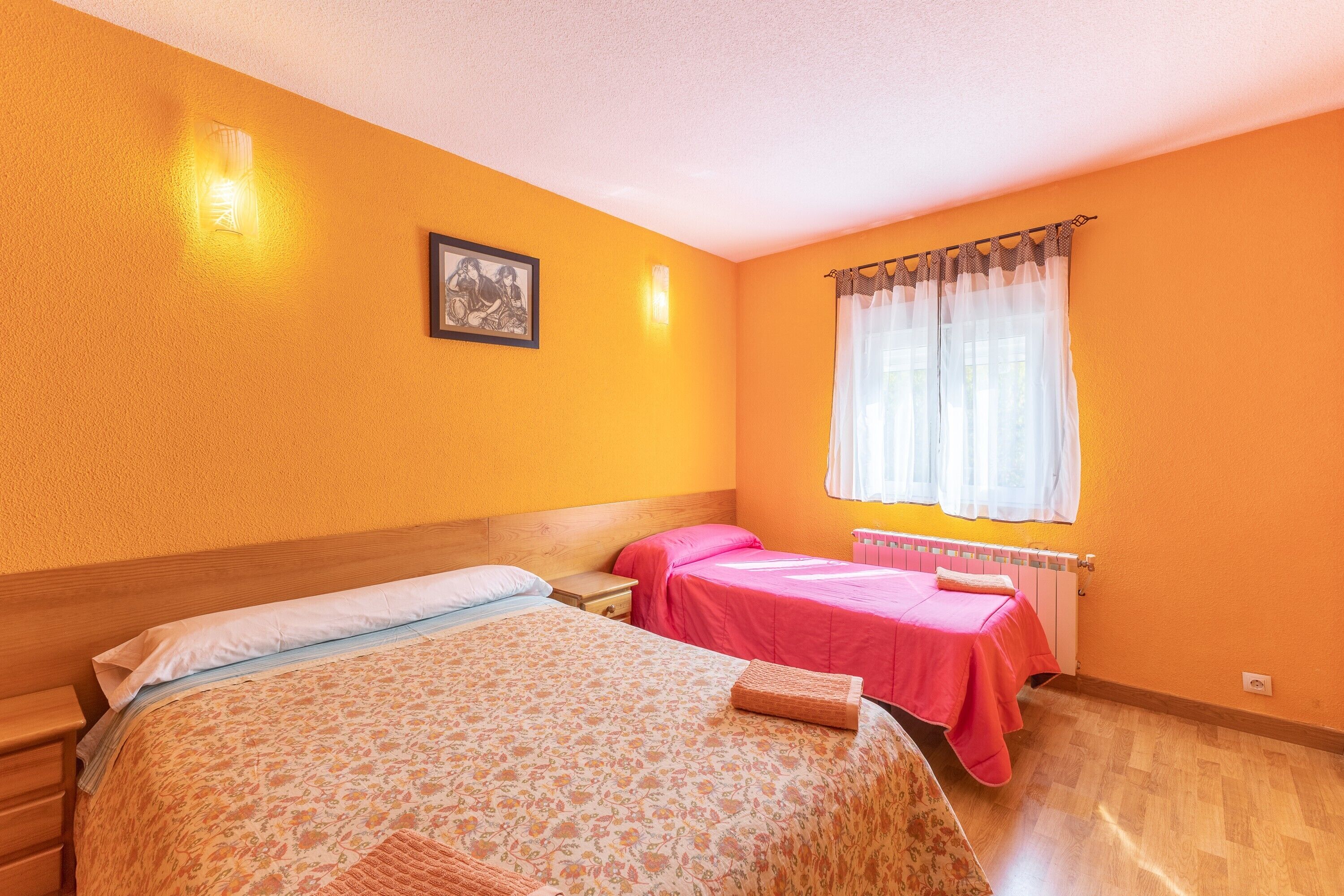 Habitación con dos camas, cabecera de madera, ventana con cortinas traslúcidas y radiador.